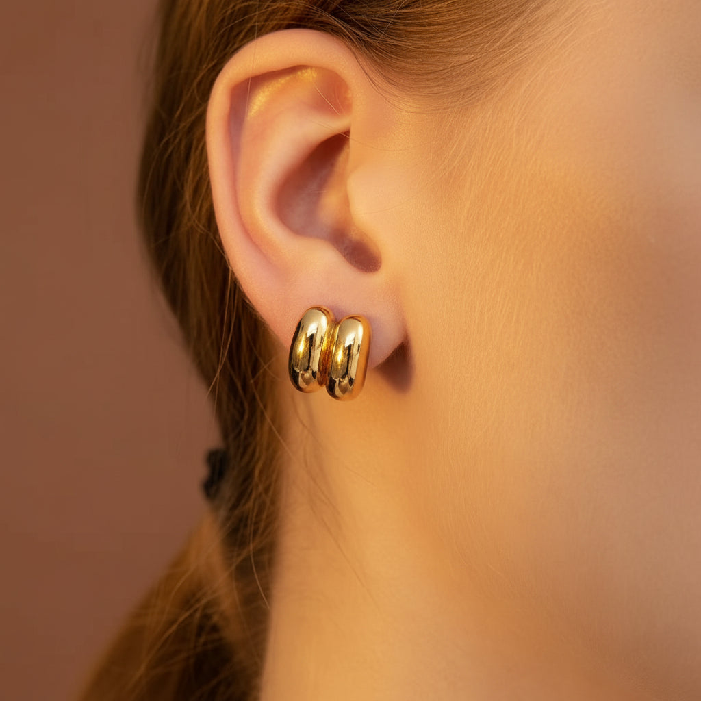 Zoomed-in lifestyle shot of a model with her face half-visible, showcasing the 18K gold plated twin-ribbed dome stud earrings against a rich brown background.