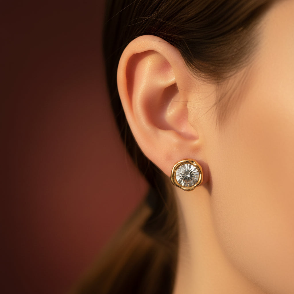 Zoomed-in lifestyle shot of a model with her face half-visible, showcasing the gold organic molten bezel solitaire stud earrings under warm golden lighting against a rich brown background.