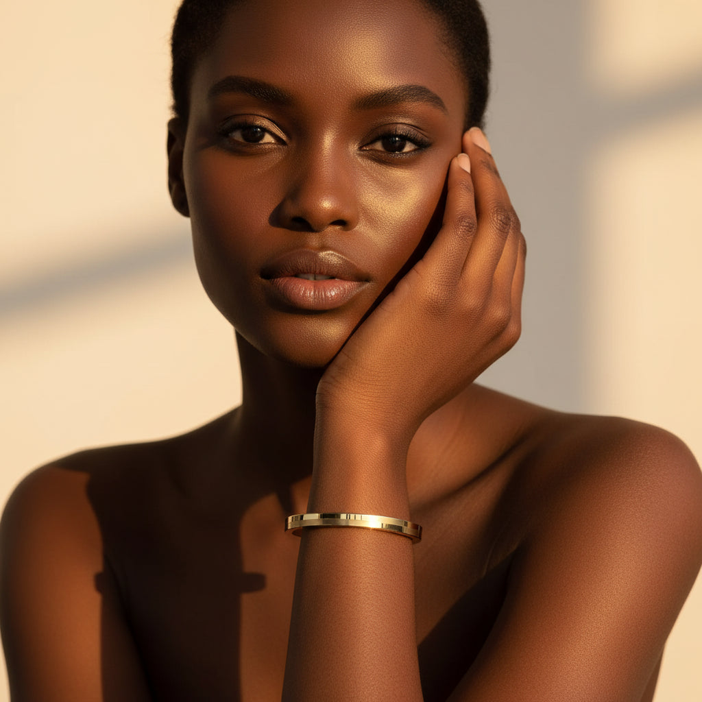 A stunning high-end e-commerce editorial photograph of a beautiful Black beauty model under warm golden lighting, with her face fully visible and the focus zoomed in tightly on the small size brick textured gold oval kada bracelet on her slender wrist.