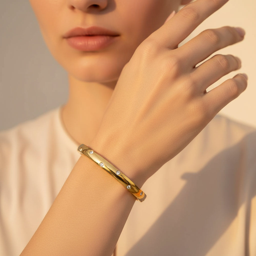 A standing profile photograph of a model (face half visible), highlighting the dainty small size gold oval kada bracelet on her thin wrist under warm golden lighting against a cream background.