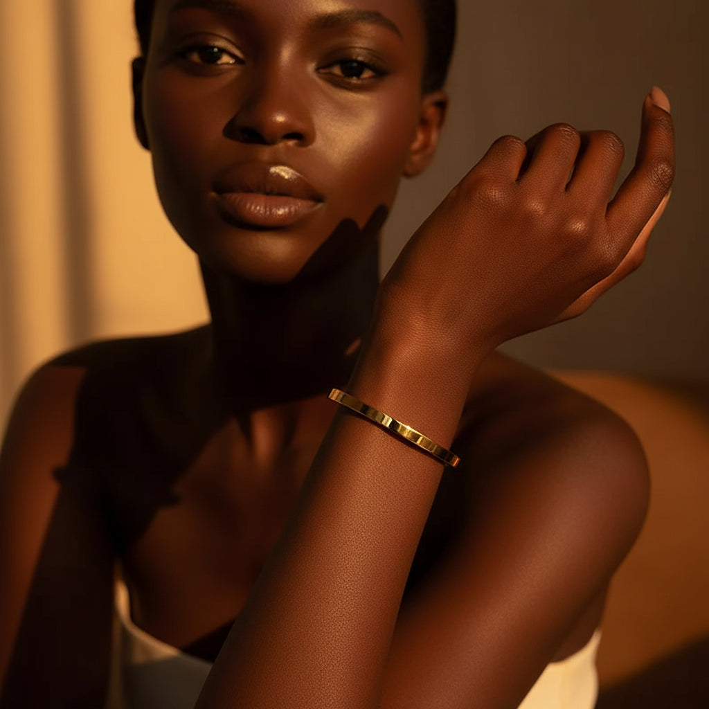 A stunning high-end e-commerce editorial photograph of a beautiful Black beauty model under warm golden lighting, with her face fully visible and the focus zoomed in tightly on the small size plain gold oval kada bracelet on her slender wrist.