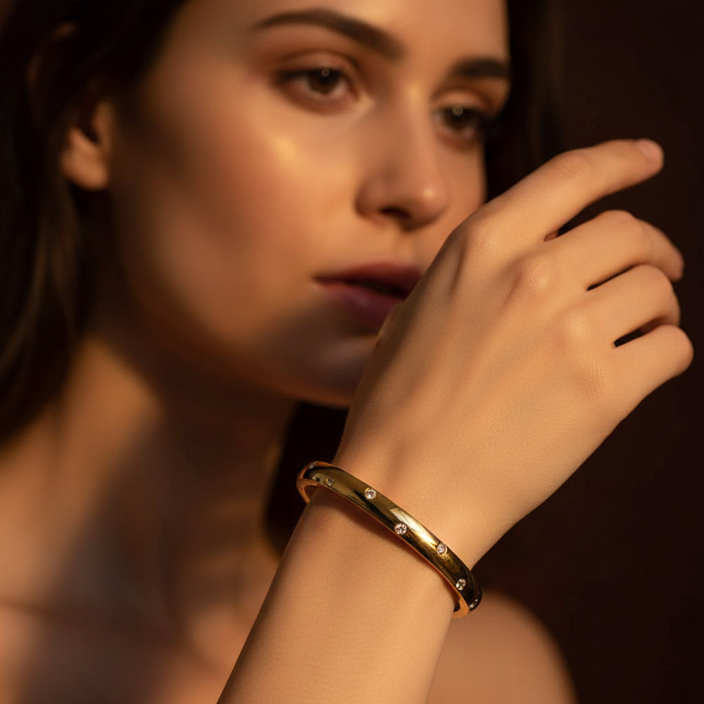A close-up flat lay photograph focusing tightly on a model's distinctly slender, thin hand and wrist wearing the small oval gold kada bangle under warm golden lighting against a deep brown background.