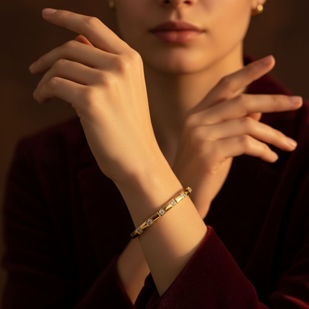 A close-up flat lay photograph focusing closely on a model's distinctly slender, thin hand and wrist wearing the small oval block-station crystal gold kada under warm golden lighting against a deep brown background.
