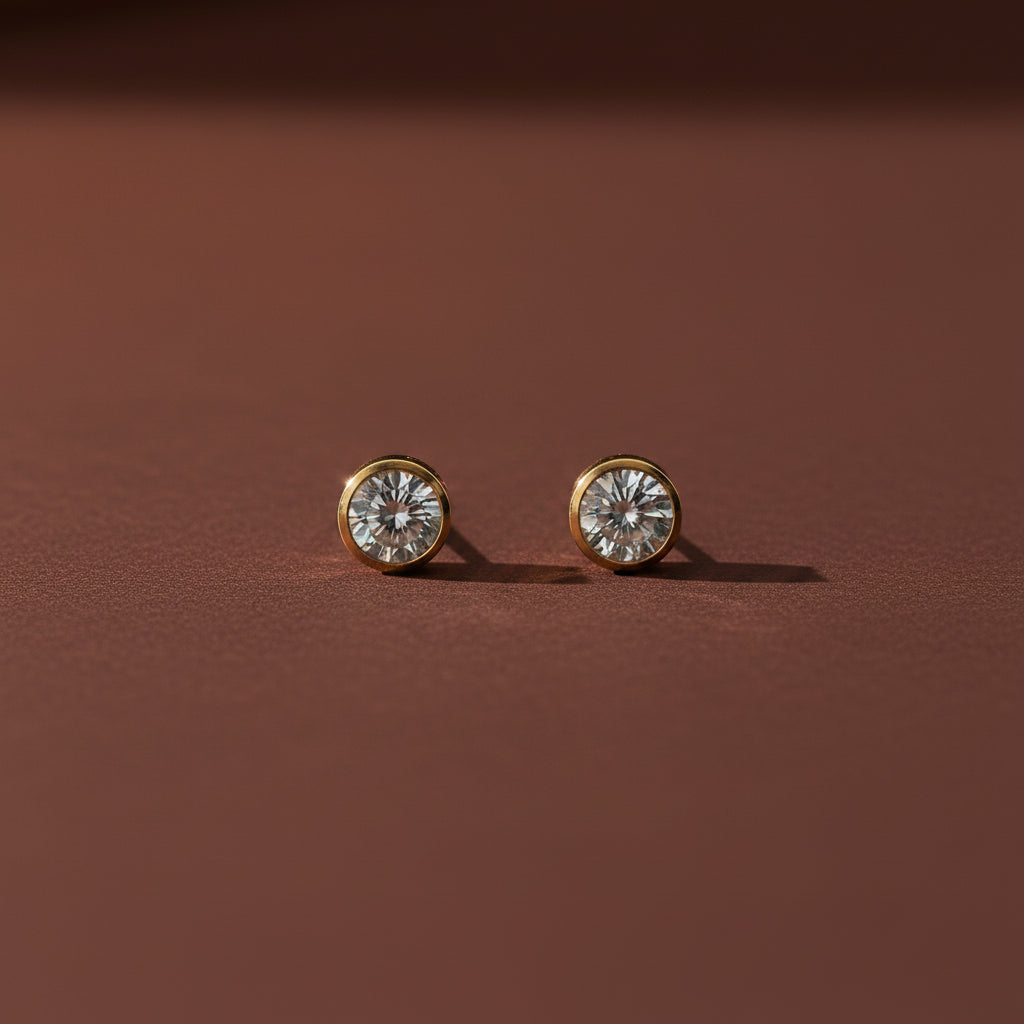 Professional catalogue flat lay of Luminary solitaire bezel stud earrings in 18K gold plating with soft shadows on a rich brown background.