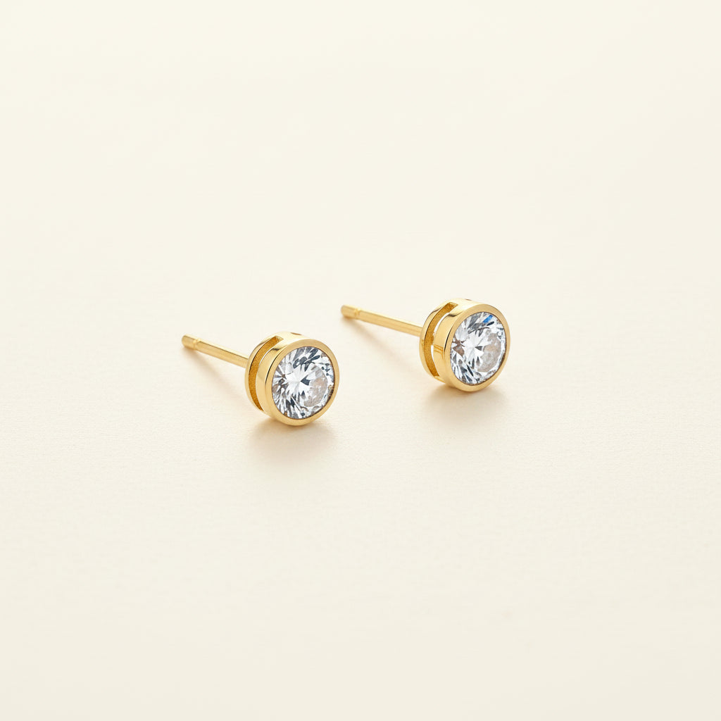 Professional standing catalogue shot of the 18K gold plated crystal bezel stud earrings with a simple shadow on a light cream background.
