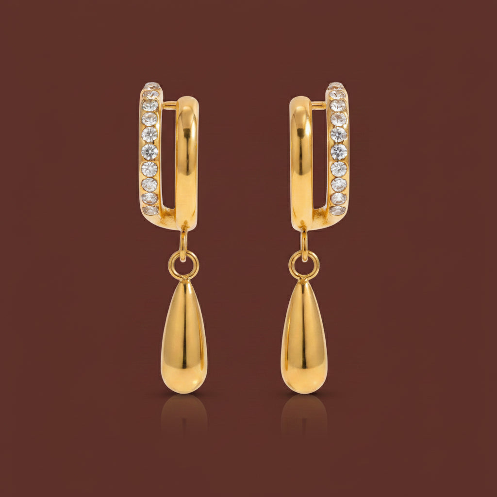 Professional catalogue flat lay of the Aura split pavé crystal teardrop drop earrings in 18K gold plating with soft shadows on a rich brown background.