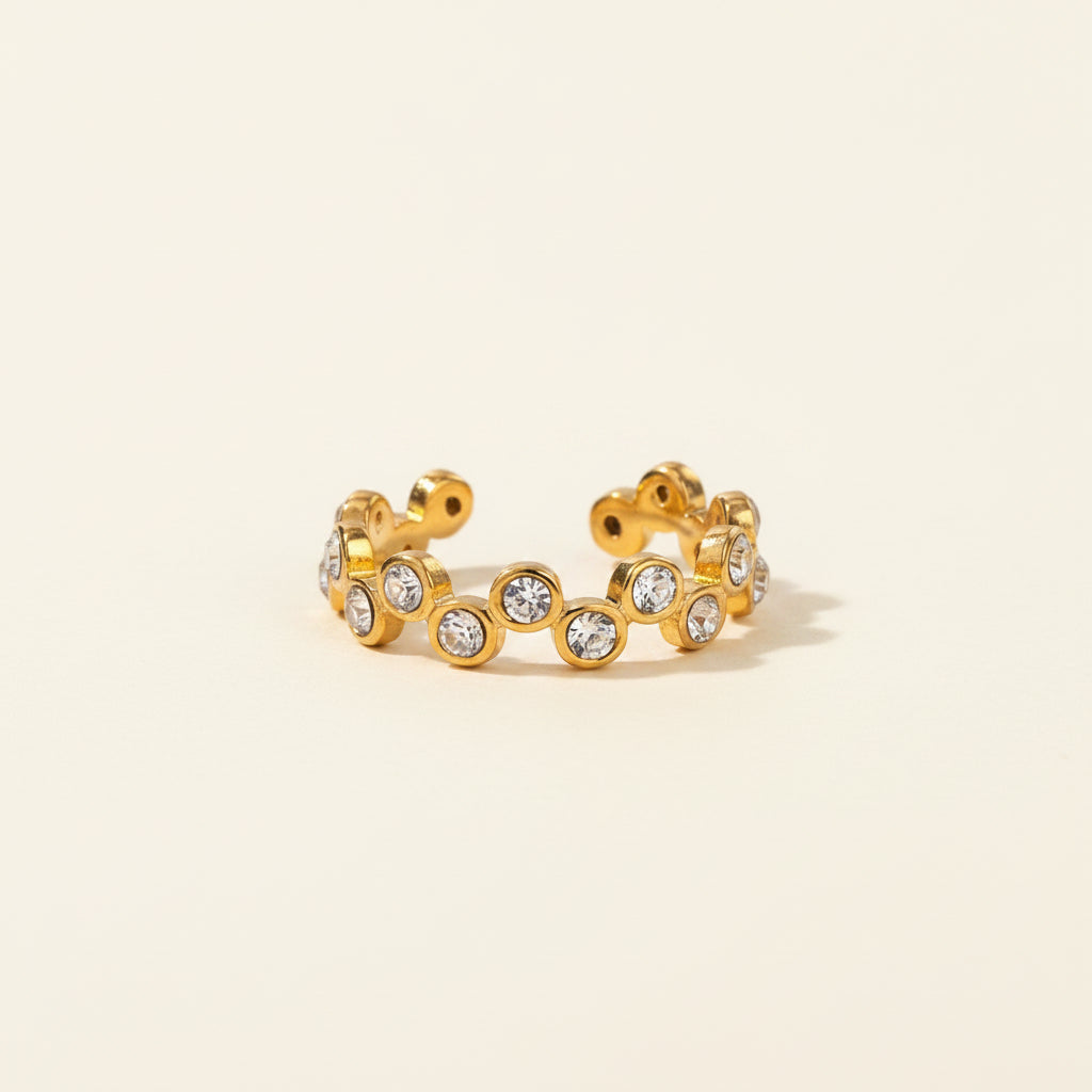 Professional standing catalogue shot of the gold plated wavy interlocking crystal bezel ring with simple shadows on a light cream background.